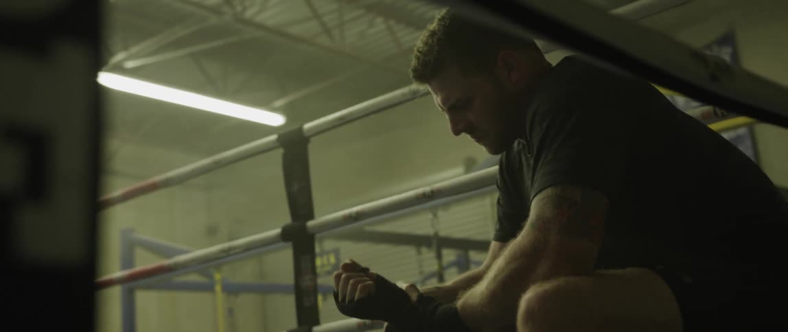 A white man sits adjusting the tape on his hands in the corner of a boxing ring in a gym