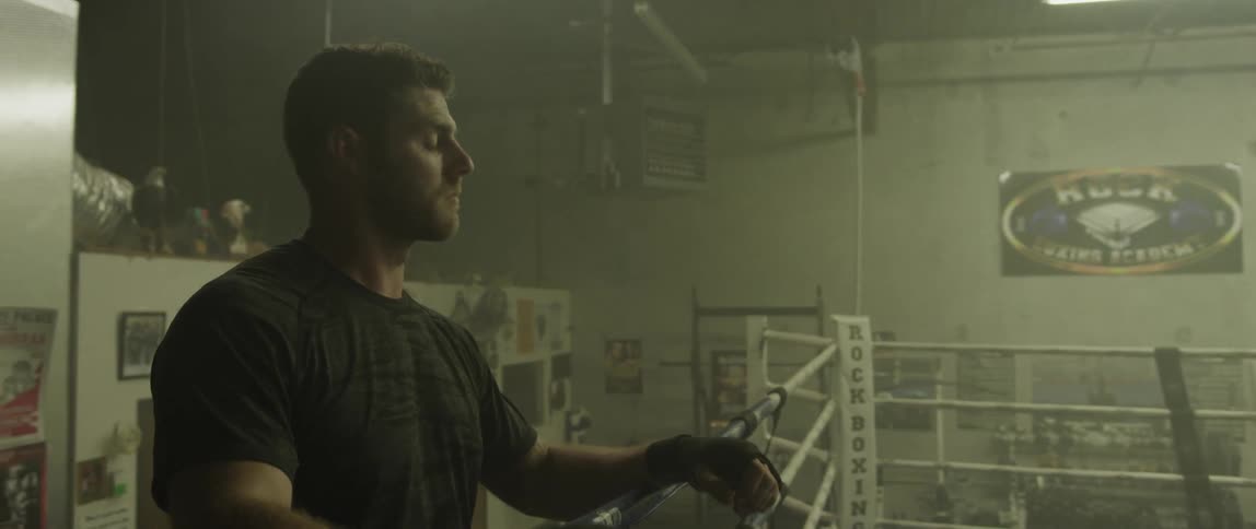 A man approaches a boxing ring in a dimly lit boxing gymnasium with a focused look on his face