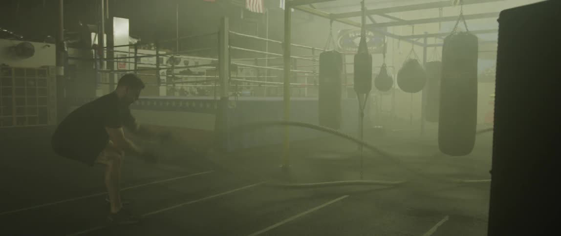 A white male uses battle ropes in a dimly lit boxing gym
