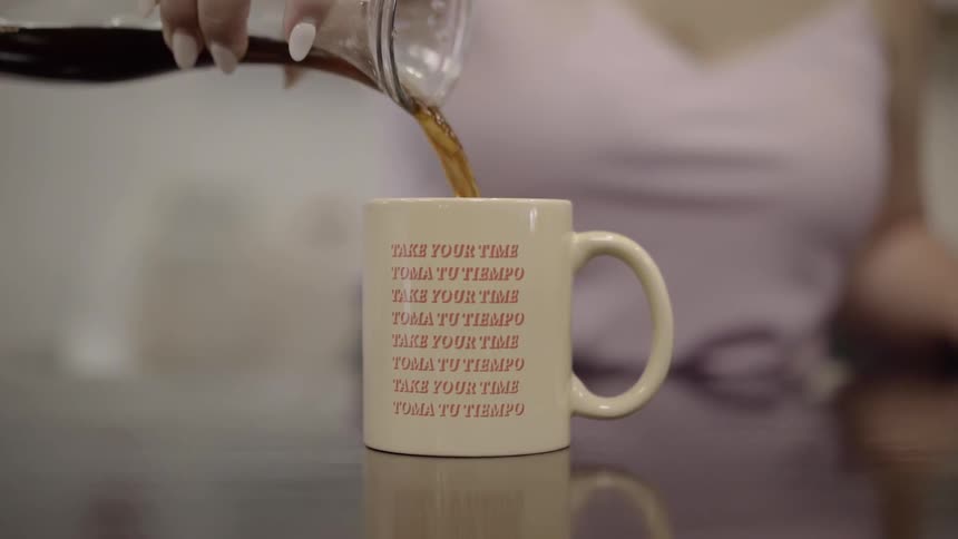 A woman is pouring coffee from a glass carafe into a tan ceramic coffee mug