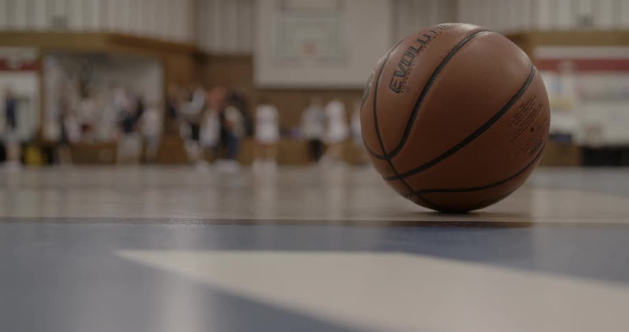 A basketball rests on the court as players shoot hoops behind it