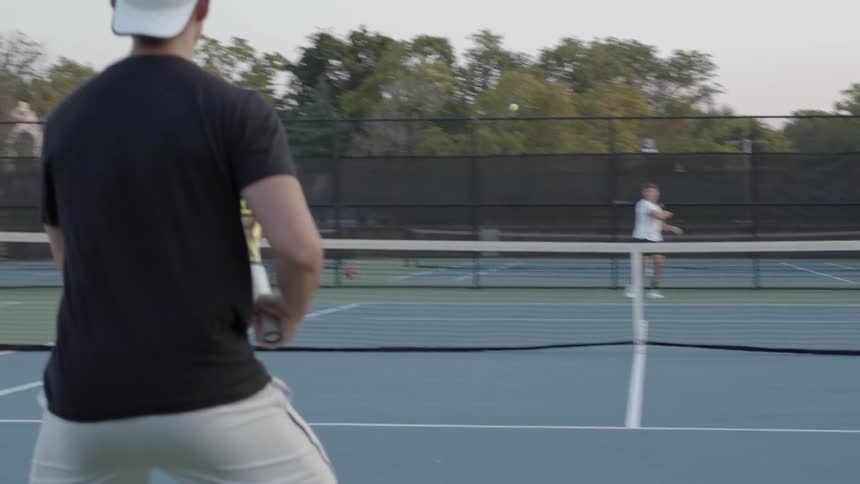 Two men play tennis on an outside tennis court in the afternoon
