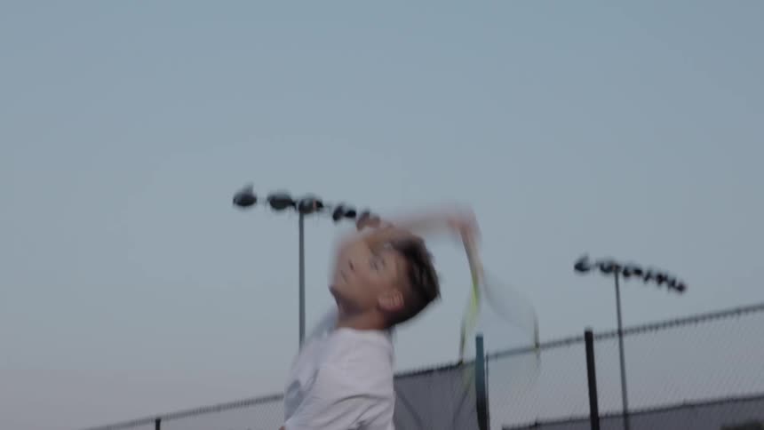 A man jumps up and hits a tennis ball at an outdoor court at sunset