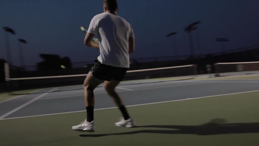 Two men play tennis on a lighted tennis court at dusk