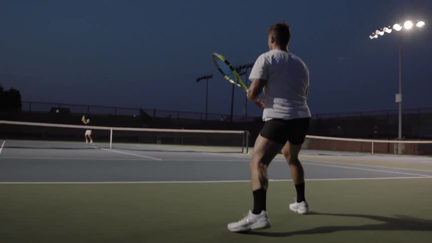 Two men play tennis on a court at night