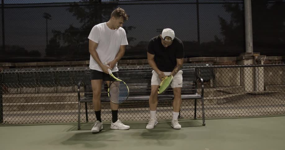 Two tennis players shake hands and sit down on bench in the tennis court at dusk