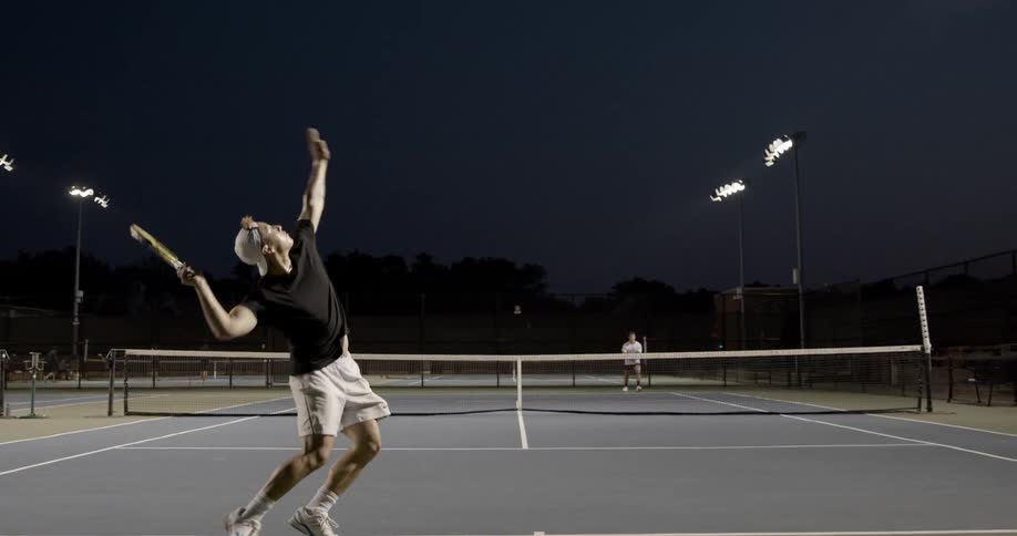 A man serves a tennis ball to his opponent in a night time tennis match on a court