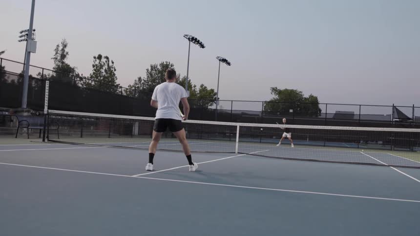 Two friends play tennis on an outdoor court at dusk