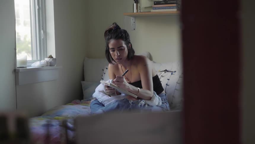 A young woman sits on a bed and writes in a notebook