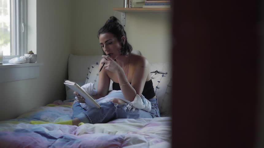 A woman is thinking sitting on her bed and then she writes on a notepad with a pen