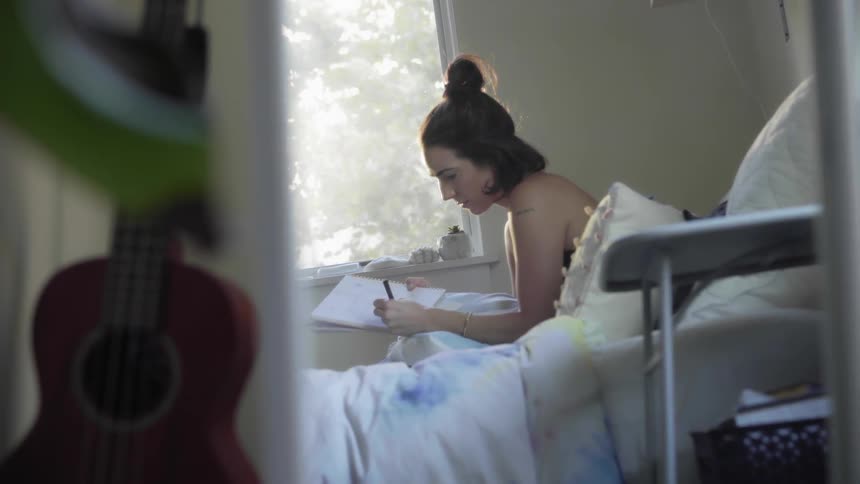 A young woman sits in her bed while writing in a notepad in daylight