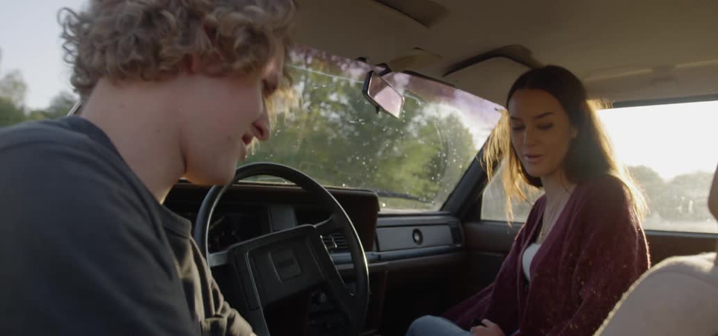 A man vacuums the driver's seat of a car while a woman sits in the passenger seat