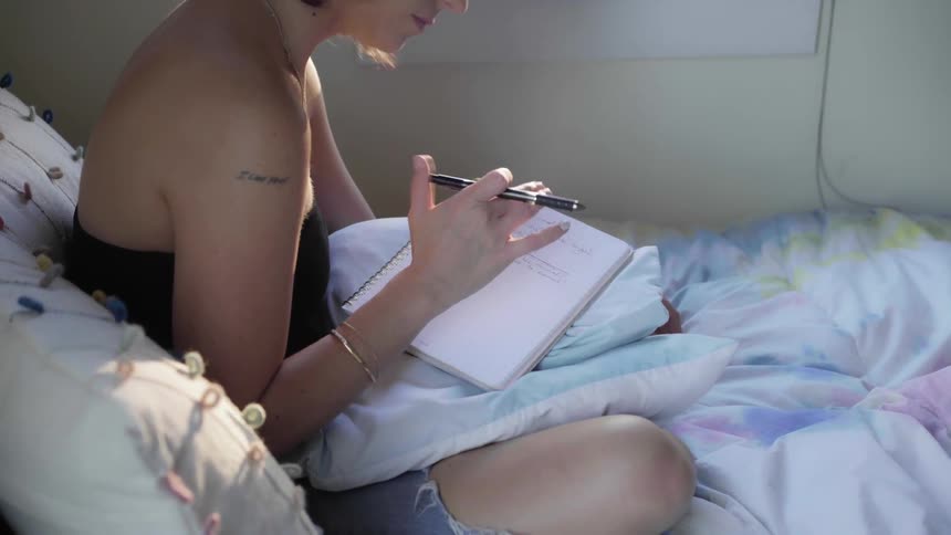 A young woman sitting on a bed moving to music while holding a pen and notebook