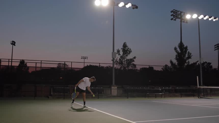 Man on tennis court at night holds racket and bounces tennis ball