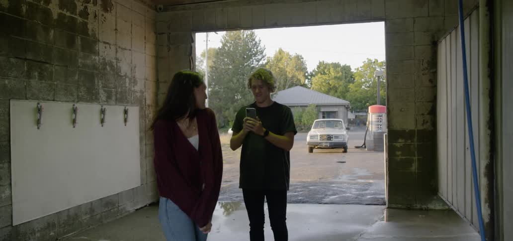 A young man holds his smartphone up to take pictures or video of his female companion who is playfully posing as they stand in an empty car wash bay at a gas station