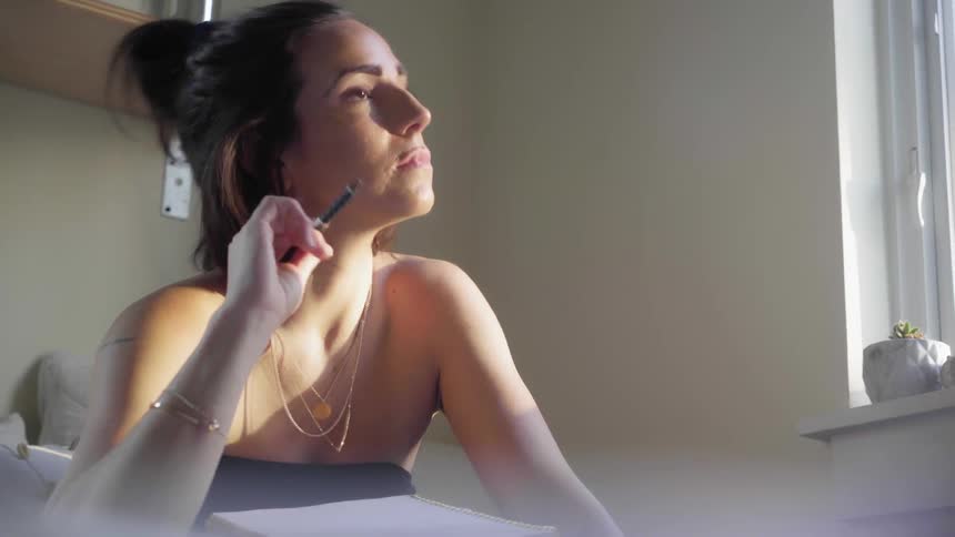A young woman wearing a tube top and gold chain necklaces looks pensively out the window of her room before she seems to decide upon what to write in her notebook with her pen