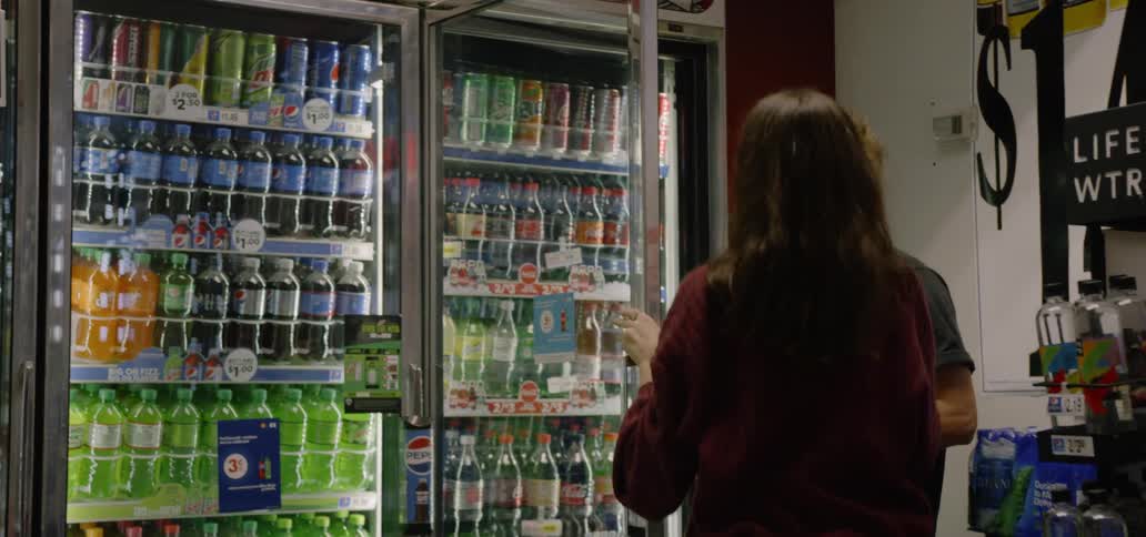 Two young people grab soda bottles from a cooler refrigerator at a grocery store
