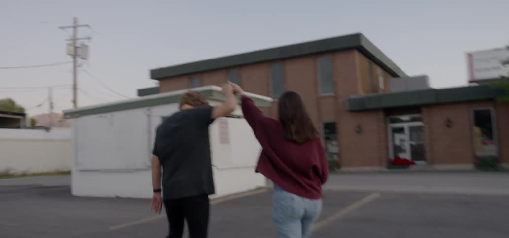A young man and woman walk through a parking lot dancing and holding hands