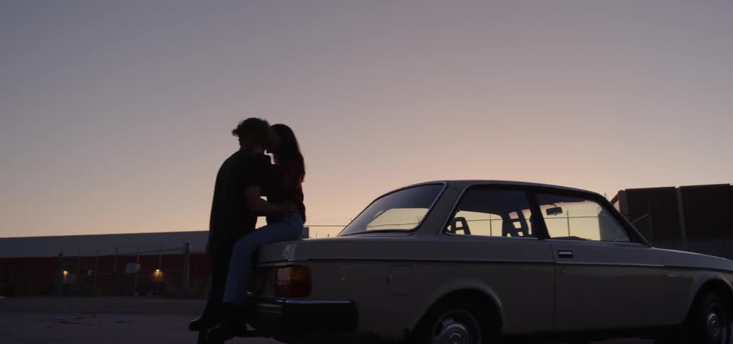 A man and woman kissing on the back of a car in a parking lot at sunset