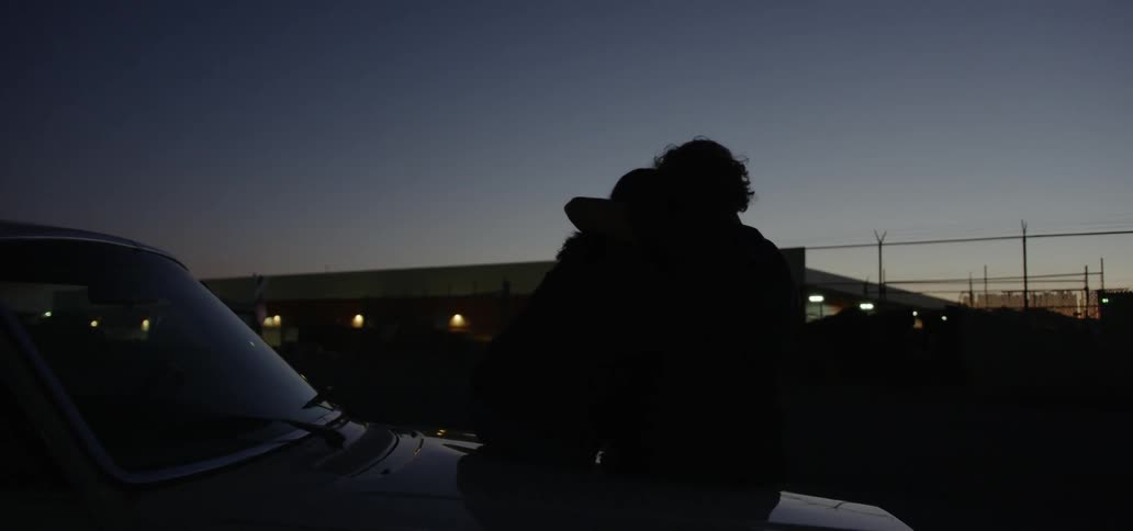 Man and woman embrace as they sit on a hood of a car at night