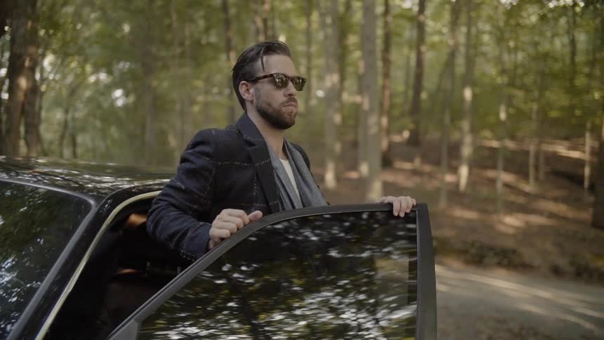 A white man wearing sunglasses gets out of a black car and then closes the car door