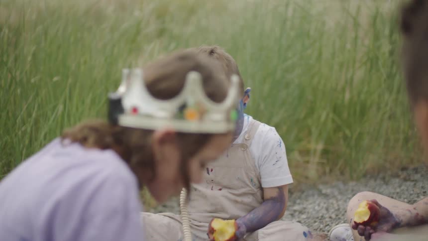 A child covered in face paint sitting outdoors eating an apple while holding a phone
