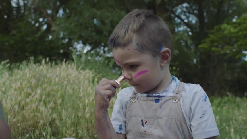 Little boy in paint splattered overall applying lipstick all over his face in a field
