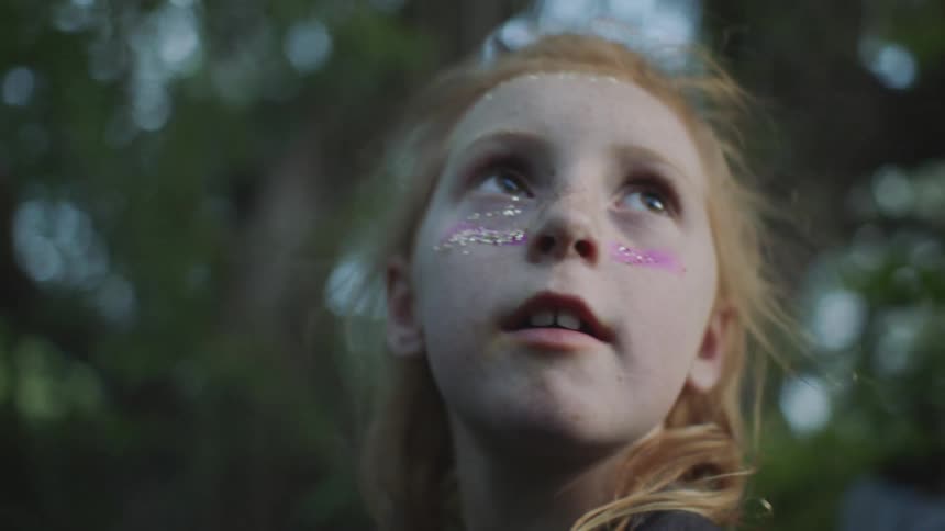 Young blonde girl with red marks under her eyes stands beside green trees blowing in the wind and looks suddenly up behind her