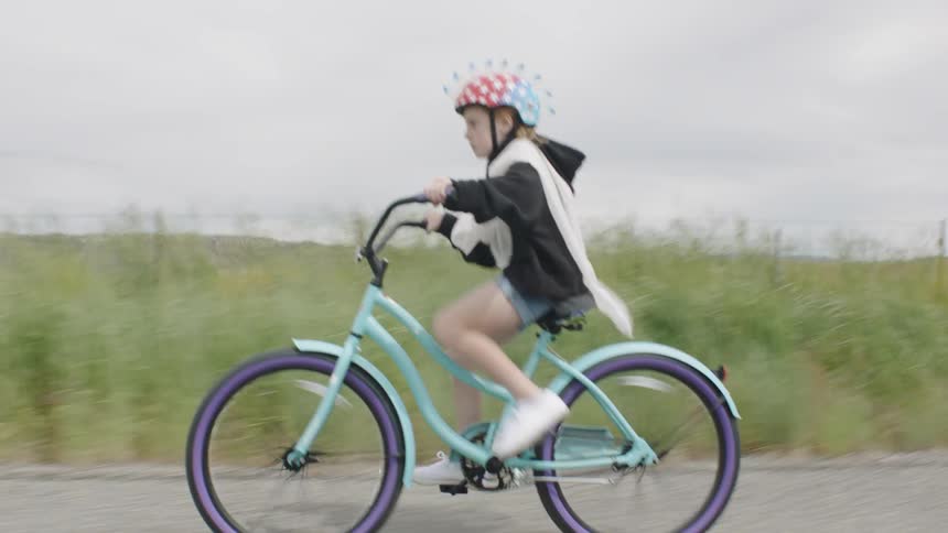 A young girl wearing a helmet with spikes rides her bike on a rural road on a sunny day