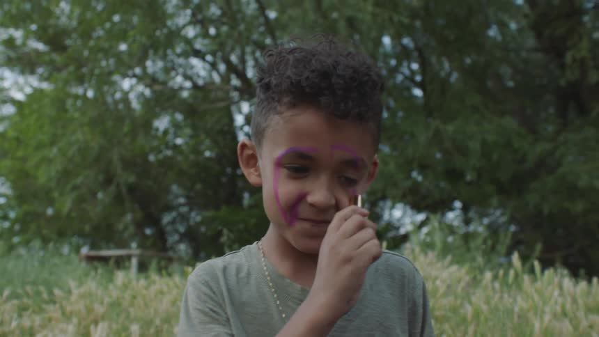Child sitting on the grass and applying purple lipstick on his face