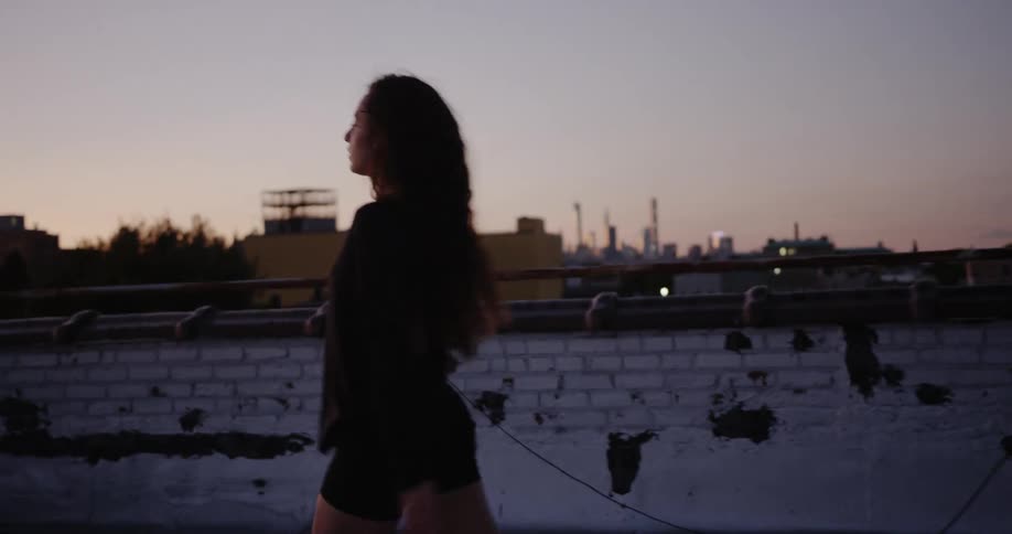 A young brunette white woman with long hair spins and leaps on a buildings rooftop