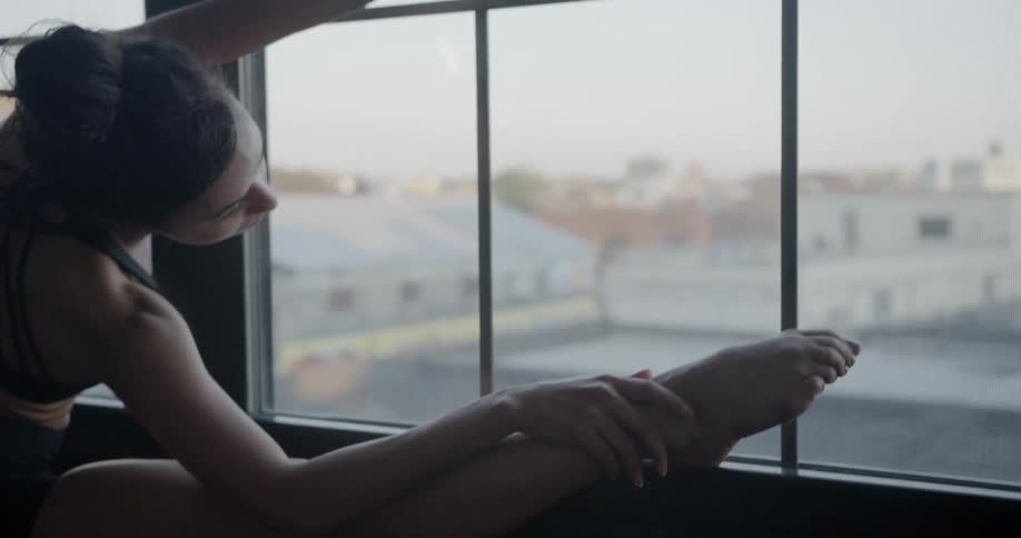 A ballet dancer stretches over her leg on a bar in front of large windows