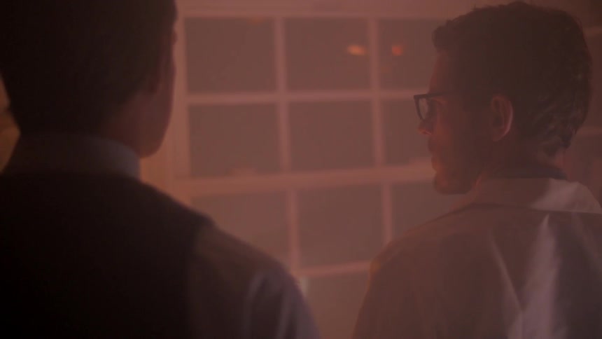 Two men stand solemnly in front of a window