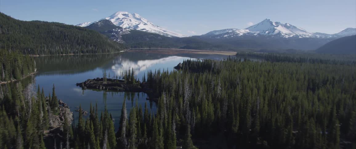 Pine trees and snowy mountains surround a calm lake on a clear day