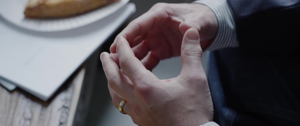 The hands of a man in a business suit and dress shirt can be seen gesturing while seated at a table with some food on it