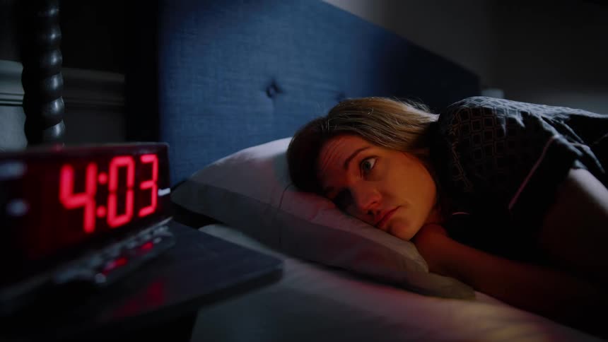 A woman laying awake in bed, looking at her clock, unable to sleep