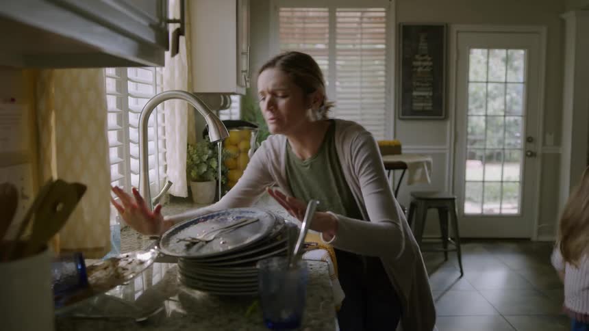 Mother washes dishes in sink and child runs up and knocks over dirty dishes