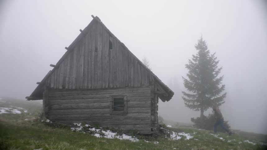 Person approaches and inspects abandoned rustic cabin in fog