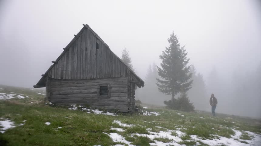 A small building made of logs sits on the side of a fog covered hill as a tall man in a thick coat approaches the building and begins to inspect its roof
