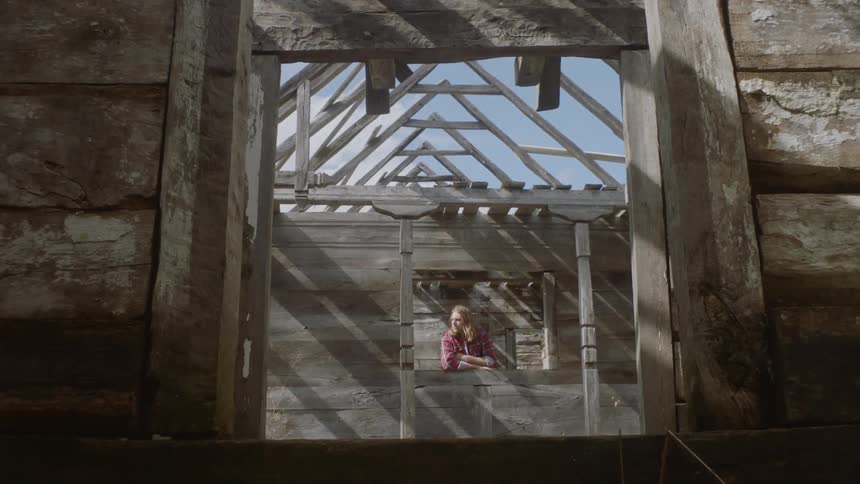 A white man leans through a window frame inside an unfinished wooden house