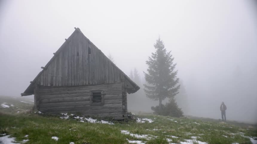 A man stands in the fog outside of a wooden cabin