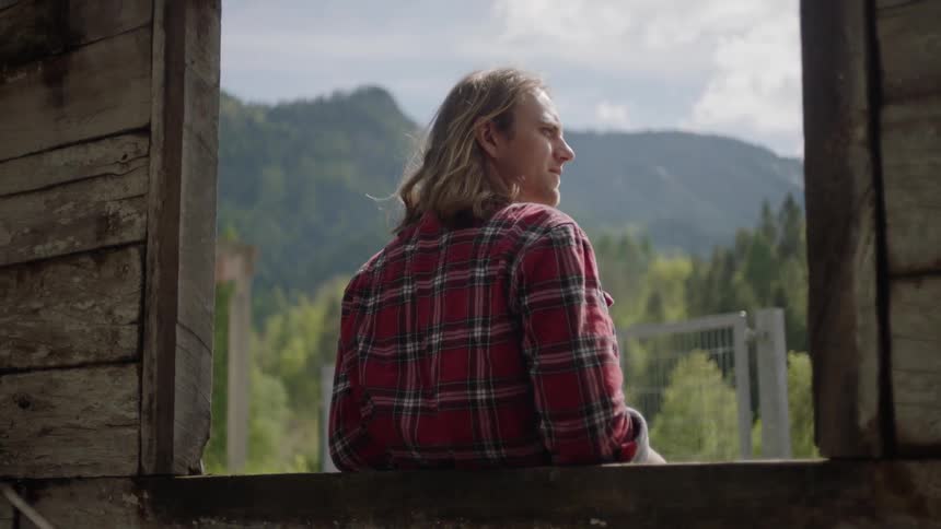 Blonde long hair man leans back on window with the landscape of the mountain during daylight