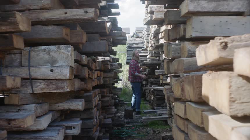 A man is walking past stacks of wooden beams