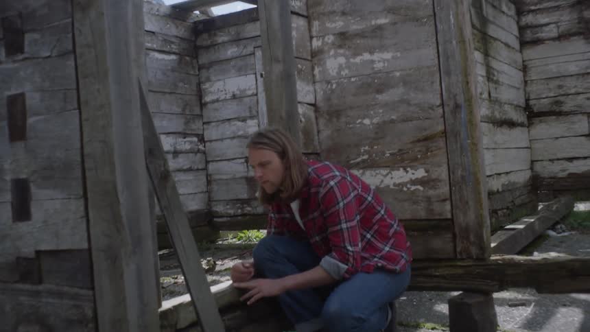 Blonde man wearing striped shirt crouches in the corner of ruined shack