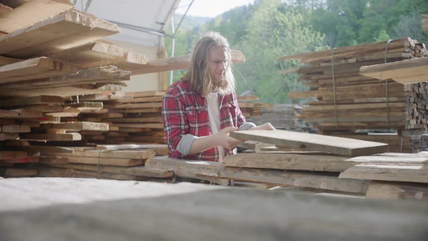 A man surrounded by stacked lumber picks up a piece of wood inspects it and tosses it down