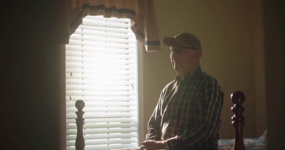 Elderly man sits on end of bed in front of window in sunlight