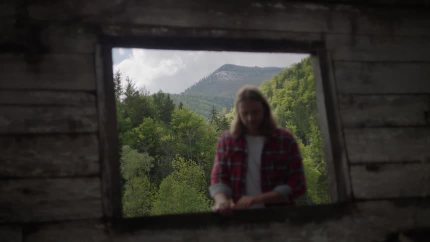 A man stands in the window of an old building with mountains behind