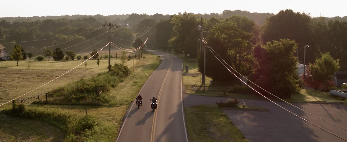 Two motorcycles traveling on a highway going through a small town near farms