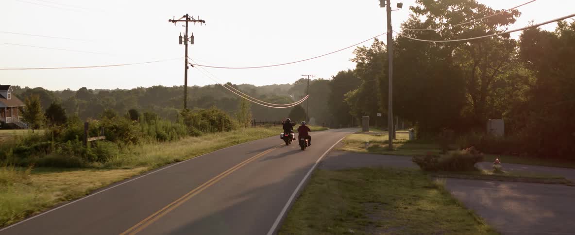 Two men ride down a two lane road through the countryside each on their own motorcycle