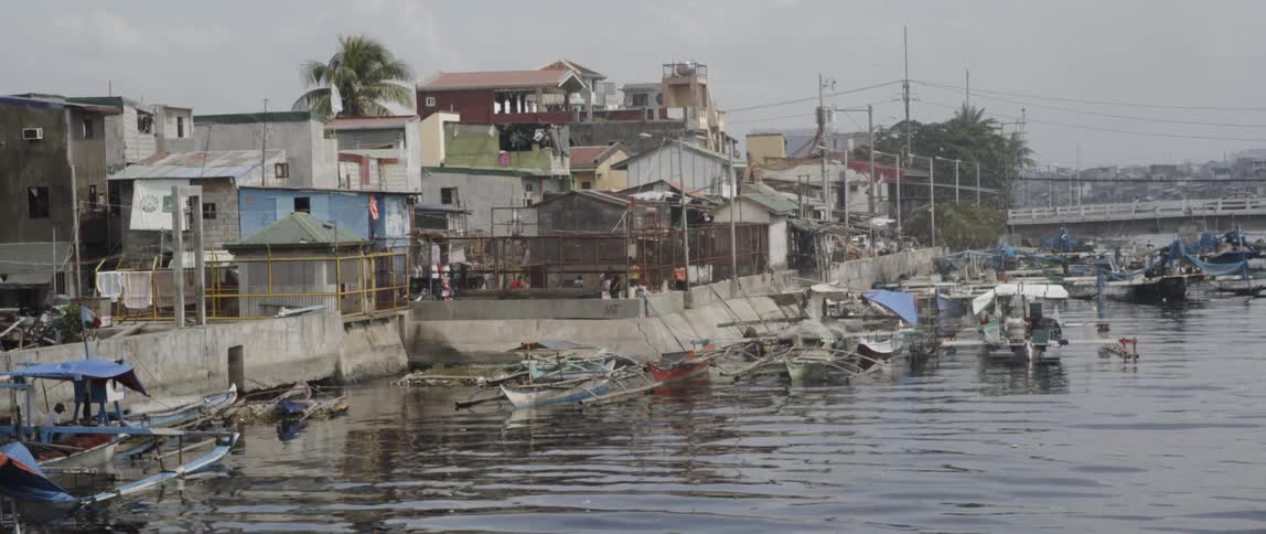 A variety of boats and canoes along a river surrounded by structures